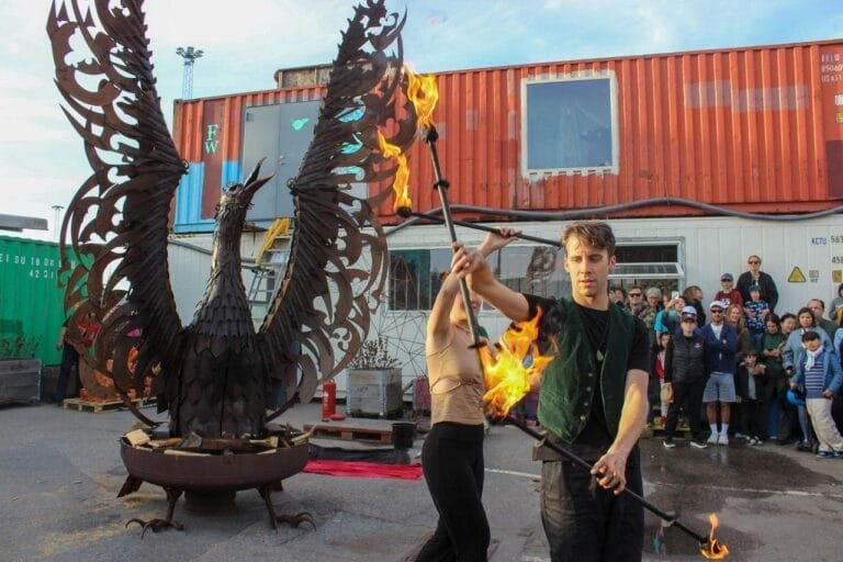 Performers lighting a large metal phoenix sculpture set over a fire basin at a Valborg street party, with a crowd watching